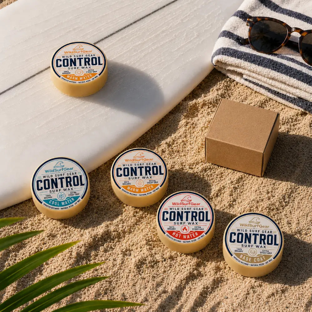 Organic surf wax tins displayed on beach sand beside a surfboard, towel, sunglasses, and kraft box